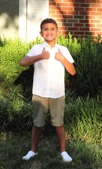 A young boy with curly brown hair stands in a yard giving a thumbs up. He is wearing a white button-down shirt and khaki shorts.