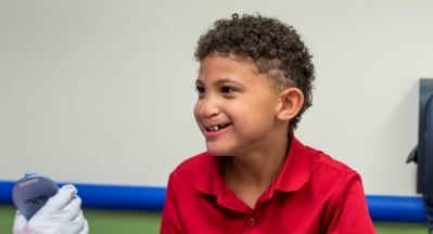 A boy with brown skin and brown curly hair smiles at a clinician who is not pictured.