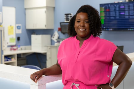 Register nurse chelsea krivanek in hot pink scrub top and pants posing against nursing station counter 