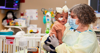 Children's Mercy provider wearing PPE and holding a toddler patient in her arms who has a tube in her nose.