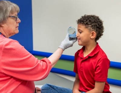 A young boy with brown skin and brown curly hair has a measuring device held up to his face in a clinic setting.