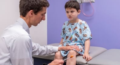 A young boy in a hospital gown gets a reflex test from a male clinician in a white shirt.