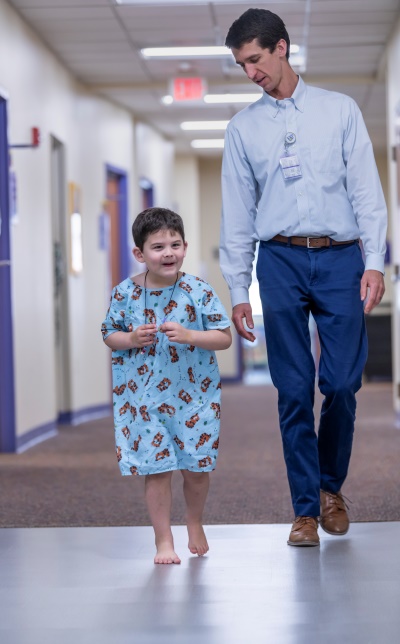 A young boy in a hospital gown walks through a hallway with a physician.