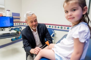 A young girl in a hospital gown sits on a clinic table while a physician tests her knee reflexes.