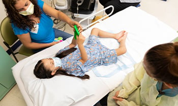 A girl in a hospital gown laying on an examination bed at Children's Mercy. The bottoms of her feet touching and knees bent out to the sides. Two Children's Mercy providers are on either side of the bed.