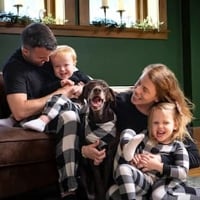 Mallory Schuh with her two children, husband, and dog wearing match pajamas. Family wearing black shirt and black and white plaid pajama pants.