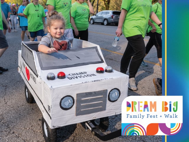 A young girl rides in a small car designed to look like an ambulance. Text reads: Dream Big Family Fest + Walk.