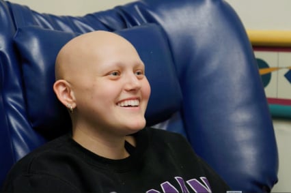 A teenage girl without hair smiles while sitting in a blue chair.