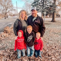 Kallie Kirk with husband and three sons posing outside in fall leaves. Kallie in green sweater and black vest, Husband and middle child in Black pull sweaters. Son on left and son on right wearing red hoodies. 