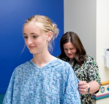 A teenage girl with blond hair wearing a hospital gown has her back measured by a clinician. 