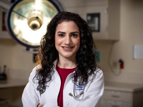 Samira Naime, MD in a physicians' coat with a large medical lamp behind her.