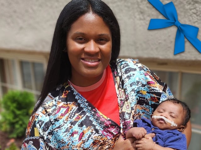 A Black woman holds a newborn boy. The boy has dark hair and tape across his upper lip.