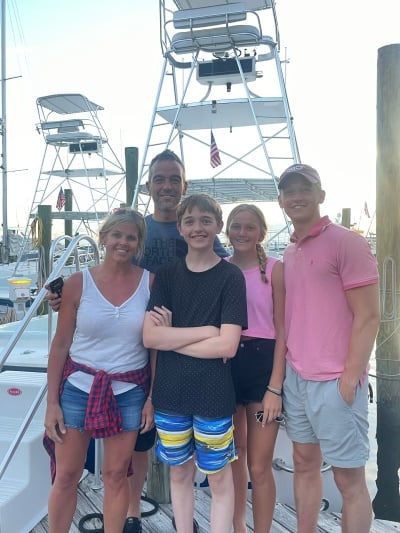 A family of 5 -- mom, dad, two boys and a girl -- stand on a docf near large boats.