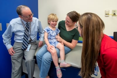 A young girl sits on a caregiver's lap on an exam table, and smiles at a woman with long brown hair. A man in a blue tie also sits on the exam table.