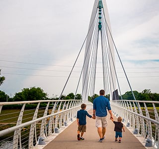 Brandon Billinger holds hands with his two young sons walking away as the cross a walking bridge on a sunny day.