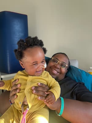 A baby laughs while being held by his dad, who is also smiling. The baby is wearing a yellow onesie and has a small tube in his nose and two tubes going to his torso. The dad has a hospital bracelet on his arm. 