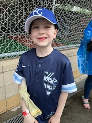 A young boy smiles while wearing a Kansas City Royals jersey and an oversized baseball cap. 