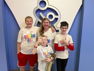 A mom and her three sons wear matching t-shirts and pose in front of a Children’s Mercy Kansas City logo sign. The t-shirts read “Dream Big,” and two of the boys are giving the thumbs up sign. 