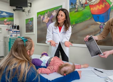 Dr. Lisa Berglund looks at X-rays while June, age 2, lays on an exam table wearing a spica cast on her legs and hips. June's mom listens to Dr. Berglund.