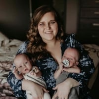 Mom, Brianna in navy blue dress sitting on the edge of a bed holding her two newborn babies. One baby in cream onesie and onw baby in grey onesie. 