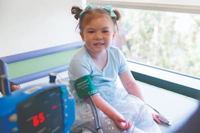 Mayia smiling, siting on exam table wearing light blue dress with arm cuff on