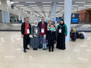 A happy family of Middle Eastern descent in traveling clothes poses for a group photo near a baggage claim in an airport. The family includes two parents, three children, a grandma and an uncle.