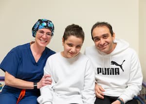 A female doctor wearing blue scrubs and a black and blue surgical cap hugs the arm of a 12-year-old boy with short brown hair wearing a white sweatshirt. The boy’s father sits on the other side of him. He also has brown hair and a white sweatshirt with a Puma logo.