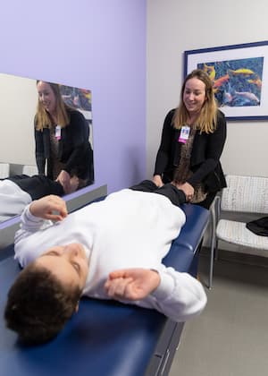 A female nurse practitioner with long, dark blonde hair is wearing a black sweater. She gently stretches out her patient’s legs. The patient is a 12-year-old boy wearing a white sweatshirt who is laying on a blue exam table. There is a mirror next to the table that reflects the boy and the nurse.