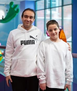 A man with brown hair and a white Puma sweatshirt stands with his arm around his son, a 12-year-old boy with brown hair and a white sweatshirt. They are standing near a purple handrail overlooking the main lobby of Children’s Mercy Adele Hall Campus. There are large, colorful sculptures hanging from the ceiling in the background.