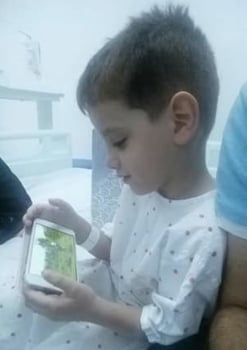 A boy about 4 years old wears a white hospital gown with pink dots. He is sitting on a bed by his father and watching a video on a cell phone.