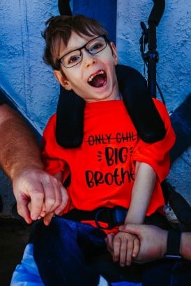 Samuel Burroughs sitting in a wheelchair and smiling. He is wearing a shirt that has the words "Only Child" crossed out and below that it reads, "Big Brother."